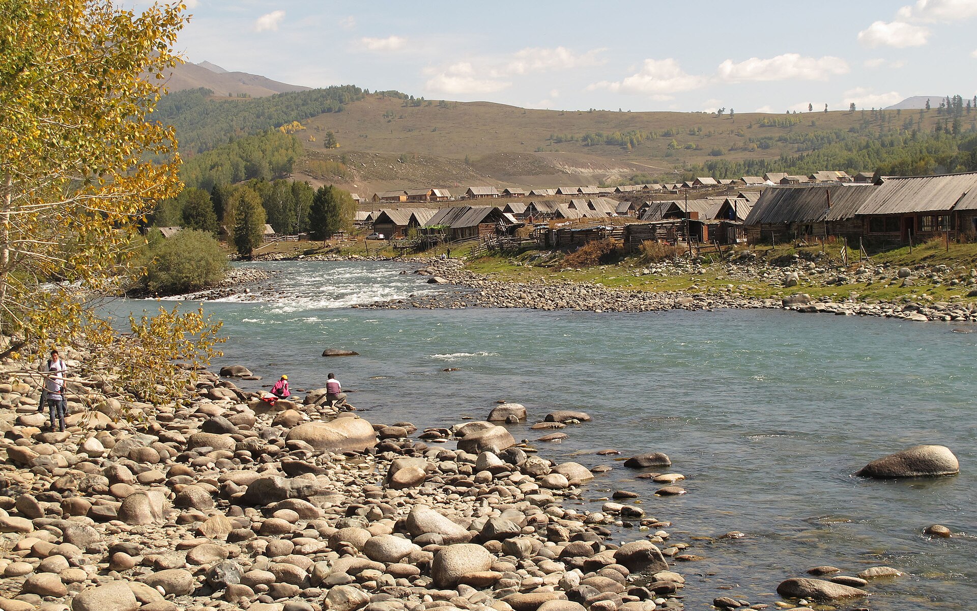 The Hemu River flows past autumn trees and a wooden-house settlement, with adults on the public riverbank.