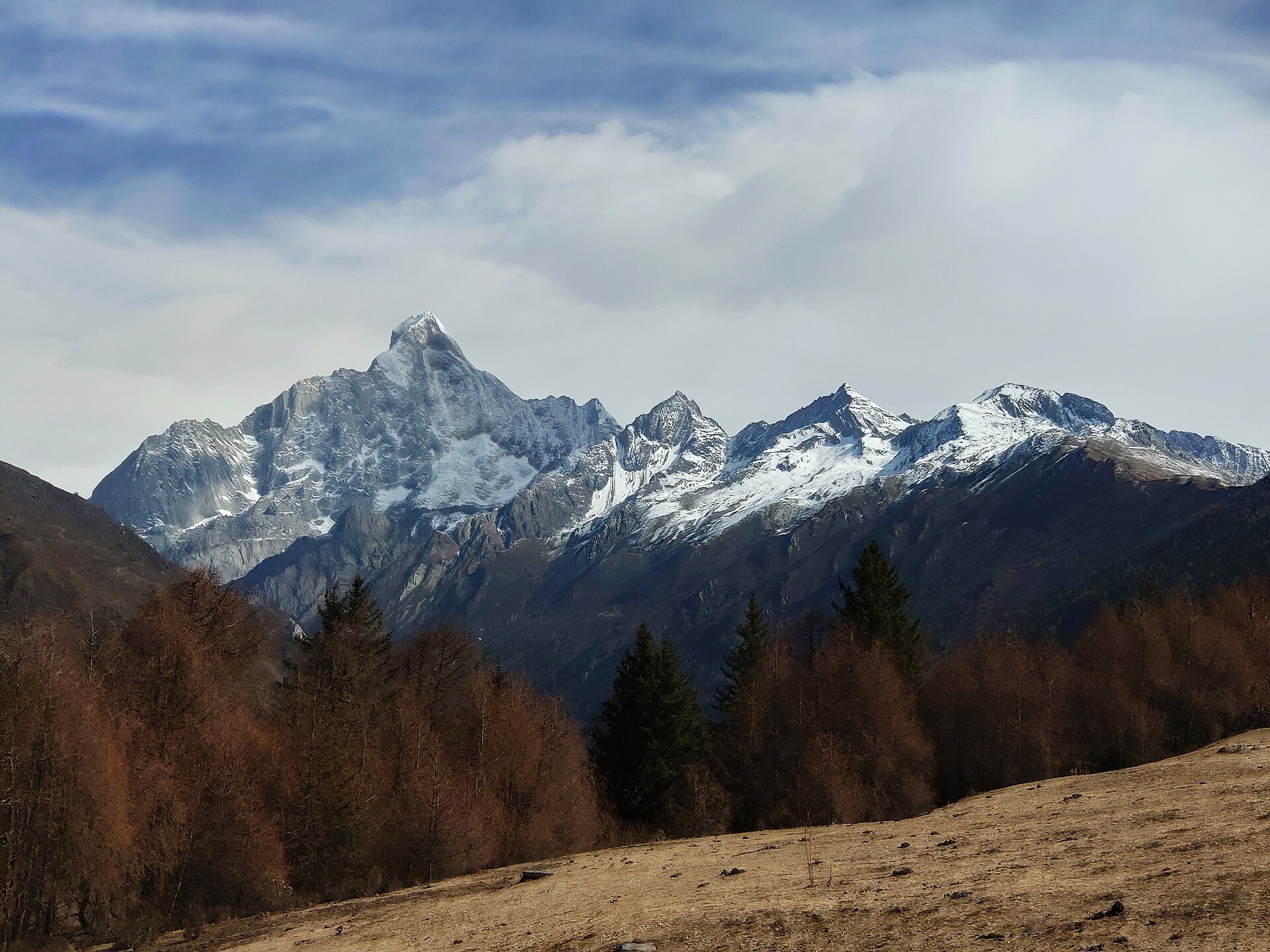 Snow-covered peaks of the Mount Siguniang group rise beyond a forested valley and meadow in March 2019.