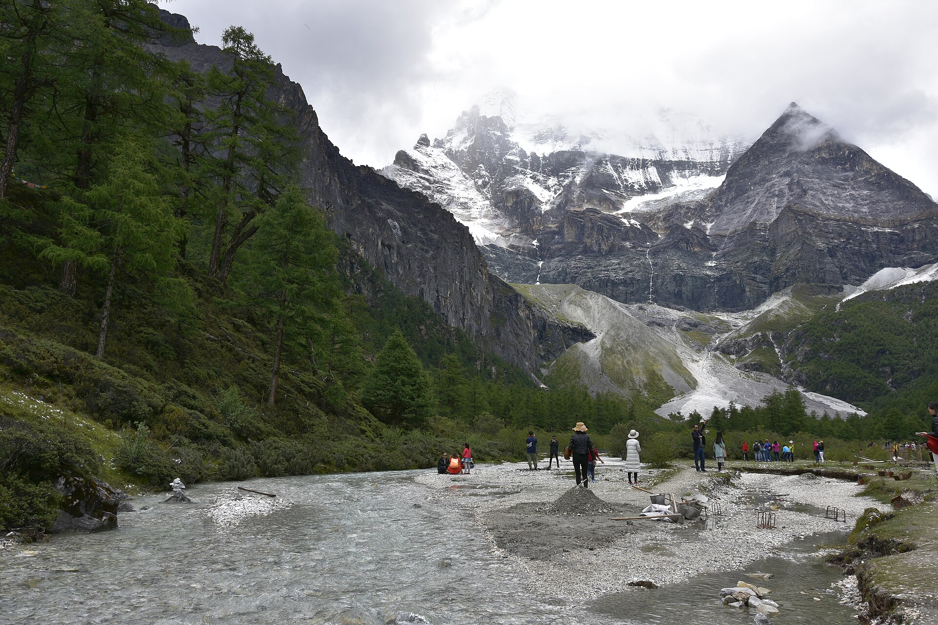 Visitors stand beside rocky shallow water at Pearl Lake below cloud-covered Xiannairi in Yading.